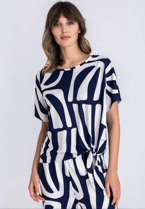 Femme portant un haut à manches courtes avec un motif abstrait bleu marine et blanc, noué sur le côté, accompagné d'un pantalon assorti, debout devant un fond uni.