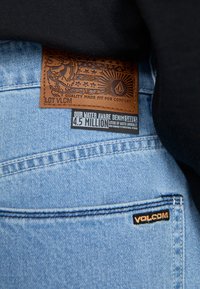Světle modré džíny z denimu mají hnědou koženou nášivku s embosovaným vzorem, logem a ekologickými informacemi na černém štítku.