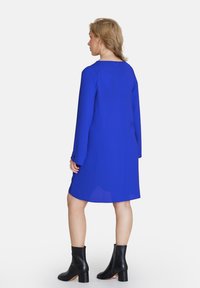 Königsblaues Kleid mit langen Ärmeln, Rundhalsausschnitt und rückseitigem Reißverschluss. Kombiniert mit schwarzen Stiefeletten mit einem blockartigen Absatz.
