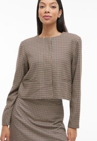 Kroćena jakna s uzorkom houndstooth u smeđoj i krem boji, s dugim rukavima, okruglim izrezom i prednjim džepovima, nošena preko odgovarajuće suknje.