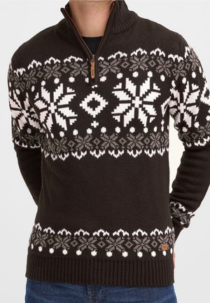 Mann trägt einen braunen Strickpullover mit weißen und grauen nordischen Schneeflockenmuster und einem Viertel-Reißverschluss-Kragen, kombiniert mit blauen Jeans.