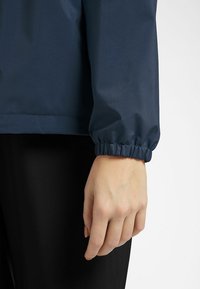 Veste imperméable marine avec poignets élastiques, texture lisse et coupe moderne. Design ajusté mettant l'accent sur le confort et la fonctionnalité.