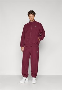 Trening burgund din material moale, compus dintr-o jachetă cu fermoar și manșete elastice, asortat cu pantaloni jogger lejeri și adidași albi.