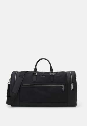 Bolsa de deporte negra hecha de tela con una textura lisa. Cuenta con dos asas, una correa para el hombro desmontable y un gran bolsillo frontal con cremallera.