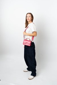 Ung kvinna med vit skjorta och mörka vida jeans, som håller en rosa axelväska, stående mot en vit bakgrund.