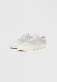 Adidași gri deschis cu superior din pânză, design cu șireturi, accente texturate în zigzag și talpă din cauciuc alb. Stil minimalistic și vârf rotunjit.