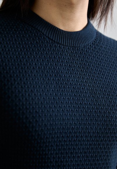 Strickpullover in dunkelblau mit strukturiertem Muster und einem gerippten Halsausschnitt, der ein geometrisches Muster über den Stoff in einer Nahaufnahme zeigt.