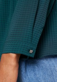 Turkos skjorta med ett strukturerat rutmönster, som har en manschett med en grå knapp dekoration. Kombinerad med denimtyg.