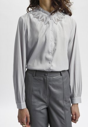 Blouse grise à manches longues avec un col brodé blanc délicat, devant à boutons, associée à un pantalon en cuir gris. Tissu doux avec un léger brillant.