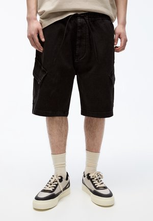 Schwarze Cargo-Shorts aus Denim mit Seitentaschen, elastischem Bund und Kordelzug. Kombiniert mit cremefarbenen, gerippten Socken und schwarz-beigen Sneakern.