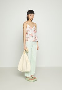 Top rosa claro, sem mangas, com padrão floral; combinado com calças largas verdes menta e sandálias chunky verdes; segurando uma mala creme.