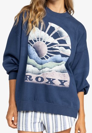 Femme portant un sweat-shirt bleu marine avec un motif soleil, montagnes, nuages et du texte "ROXY", assorti à un short rayé.