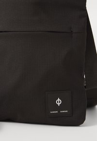 Black fabric bag with a grid texture, featuring a front pocket and a square logo patch with the brand name “SAMSØE SAMSØE.”