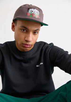 Junger Mann mit grauer Mütze mit floralem Muster und schwarzem Columbia-Sweatshirt sitzt mit neutralem Gesichtsausdruck vor einfachem Hintergrund.