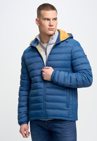 Blå polstret jakke med en gul hætte, kviltet tekstur, lynlås foran og sidelommer. Båret over en grå sweater og blå jeans.