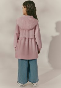 Giacca impermeabile rosa con cappuccio, vita con laccetto e polsini elastici, abbinata a pantaloni in denim a gamba larga e scarpe slip-on bianche.