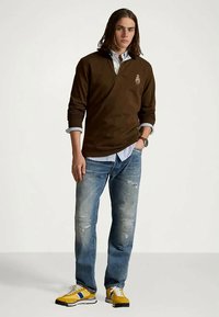 Un jeune homme portant un haut à manches longues marron sur une chemise rayée, un jean bleu déchiré et des baskets jaunes, se tenant avec une main dans la poche.