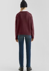 Bordeaux gebreide trui met een relaxed fit en geribbelde zoom, gecombineerd met donkerblauwe jeans en zwarte enkellaarsjes. Muts op het hoofd.
