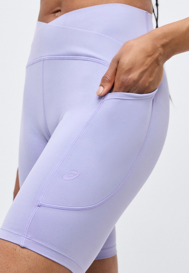 Vrouw draagt lichtpaarse, hooggetailleerde sportshorts met zijzak, hand in de zak gestoken, zichtbaar merkembleem.