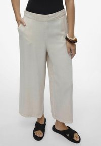 Pantaloni beige a gamba larga con tasche laterali, caratterizzati da una texture liscia e una vestibilità morbida, abbinati a sandali intrecciati neri e un bracciale multicolore.