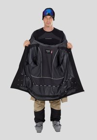 Černá lyžařská bunda s kontrastní podšívkou, která zdůrazňuje kapsy a reflexní prvky. Kombinována s béžovými lyžařskými kalhotami a lyžařskými botami.