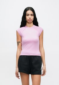 Fekete hosszú hajú nő, aki egy testhezálló, levendulaszínű, ujjatlan Nike felsőt és fekete Nike rövidnadrágot visel, egyszerű fehér háttér előtt áll.