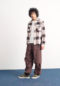 Bordeaux languotas marškos su ruda, žalia ir oranžine spalvomis, derinami su laisvomis rudomis kelnėmis ir alyvuogių žaliais sportbačiais ant mėlynos kiliminės dangos.