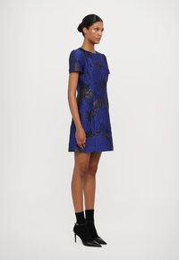 Īsām piedurknēm, teksturēta kleita dziļā zilā krāsā ar melnajiem ziedu akcentiem. Valkāta ar melnām potīšu zābaku kurpēm ar smailu purngalu un zelta aksesuāriem.