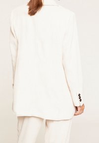 Biały żakiet z korduroyu o długim fasonie, ze stukturalnymi ramionami i czarnymi guzikami na mankietach. Teksturowana, prążkowana tkanina.