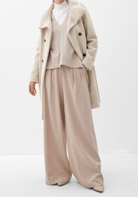 Donna con pantaloni beige a gamba larga, un gilet abbinato sopra un dolcevita bianco e un cappotto beige a doppio petto testurizzato con bottoni neri.