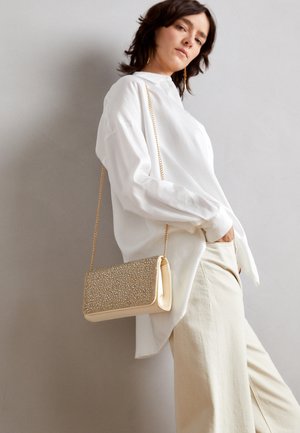 Femme portant une chemise blanche surdimensionnée et un pantalon crème, portant un sac à main beige texturé avec une bandoulière en chaîne dorée sur l'épaule.