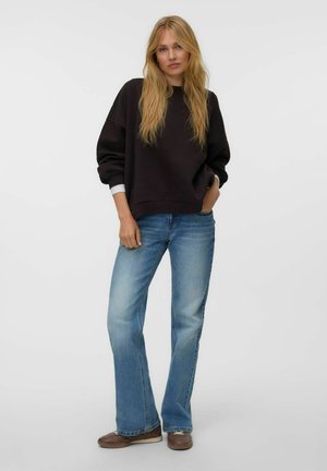 Giovane donna con lunghi capelli biondi che indossa una felpa nera oversize, jeans blu e scarpe marroni, in piedi con una mano in tasca.