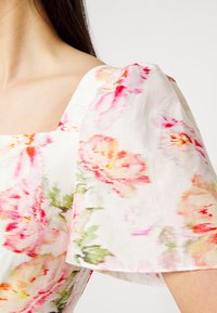 Robe blanche avec une grande manche bouffante présentant un motif floral doux rose, orange et vert sur l'épaule et le haut de la poitrine d'une femme.