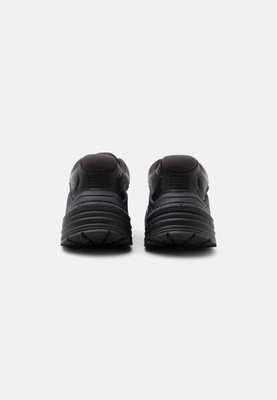 Paire de chaussures de sport noires vues de dos, avec des semelles épaisses rainurées et des collerettes de talon rembourrées sur un fond clair.