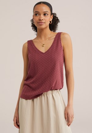 Mujer con un top burdeos sin mangas y texturizado y una falda crema, complementado con pendientes de aro de oro, un collar con colgante y anillos.