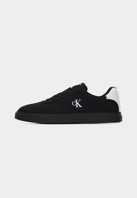 Basket basse noire avec accent blanc au talon, semelle noire et logo blanc "cK" sur le côté, montrée en profil sur fond blanc.