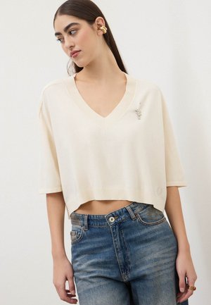 Giovane donna che indossa un maglione crema a scollo a V ampio con maniche a gomito e jeans blu a vita alta, guarda di lato.