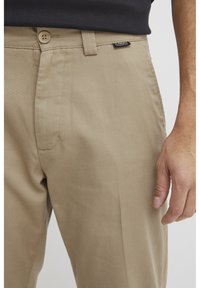 Beige Baumwollhosen mit glatter Textur, ausgestattet mit zwei Vordertaschen, einem Einzelknopfverschluss und dezentem Branding am Bund.