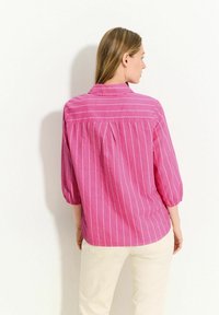 Camicia a righe rosa con maniche a tre quarti, colletto e dettagli di piega sul retro. Realizzata in tessuto leggero con una texture morbida.