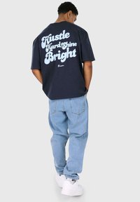 Mornarsko plava oversized majica s bijelim zakrivljenim natpisom "Hustle Hard Shine Bright" na leđima; uparena sa svijetloplavim baggy trapericama i bijelim tenisicama.