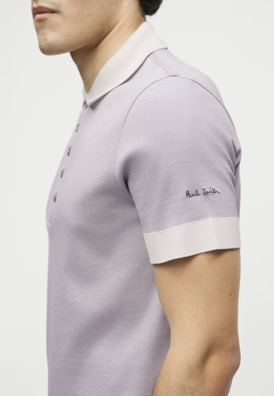 Άνδρας που φοράει ανοιχτό μοβ κοντομάνικο πόλο με λευκό γιακά και μανσέτες στα μανίκια, με κεντημένο το «Paul Smith» στο μανίκι.