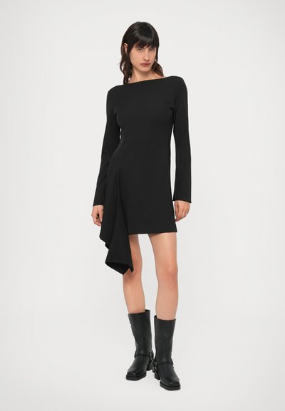 Čierne šaty s dlhým rukávom, lodičkovým výstrihom a asymetrickým lemom. Vyrobené z hladkej látky, doplnené čiernymi čižmami po kolená.