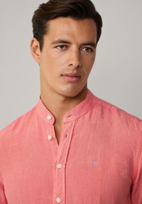 Licht koraalkleurig linnen overhemd met een mandarin kraag, knoopsluiting en een klein blauw geborduurd logo op de borst. Gladde textuur.