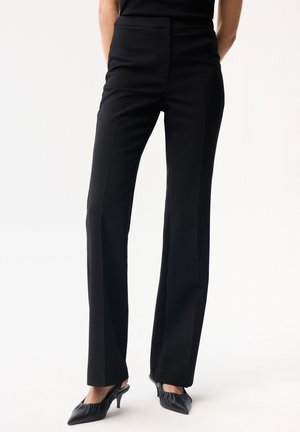 Kvinde iført højtaljede sorte bukser med lige ben og frontfold samt sorte spidse slingback-hæle.