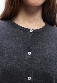 Κοντινό πλάνο ενός ατόμου που φοράει σκούρα γκρι ζακέτα με κουμπιά, με τρία εμφανή λευκά κουμπιά και μακριά κ αστανά μαλλιά.