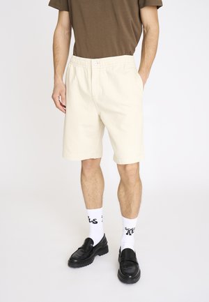 Mand iført beige knælange shorts, hvide mærkede sokker, sorte loafers og en brun kortærmet skjorte, stående med hænderne i lommerne.