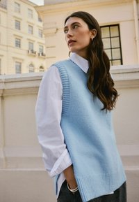 Lichtblauwe gebreide sweater-vest met textuur ribaccenten, gedragen over een wit collshirt, met een ontspannen pasvorm en zijsplitten.