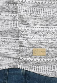 Grå og hvit mønstrete stoff med geometriske design, med et beige merke merket "SOLID" festet til den nederste sømmen, over blå denim.