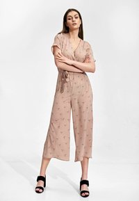 Jasnoróżowy kombinezon o kroju kopertowym, z krótkimi rękawami i wiązaniem w talii, zdobiony subtelnym wzorem ptaków i szerokimi, ściąganymi nogawkami. Czarne szpilki.