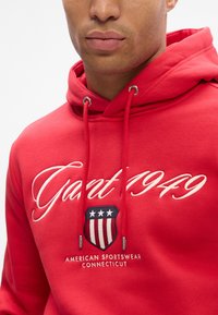 Homme portant un sweat à capuche rouge avec le texte brodé "Gant 1949" et un logo de bouclier avec des étoiles et des rayures, "American Sportswear Connecticut" en dessous.